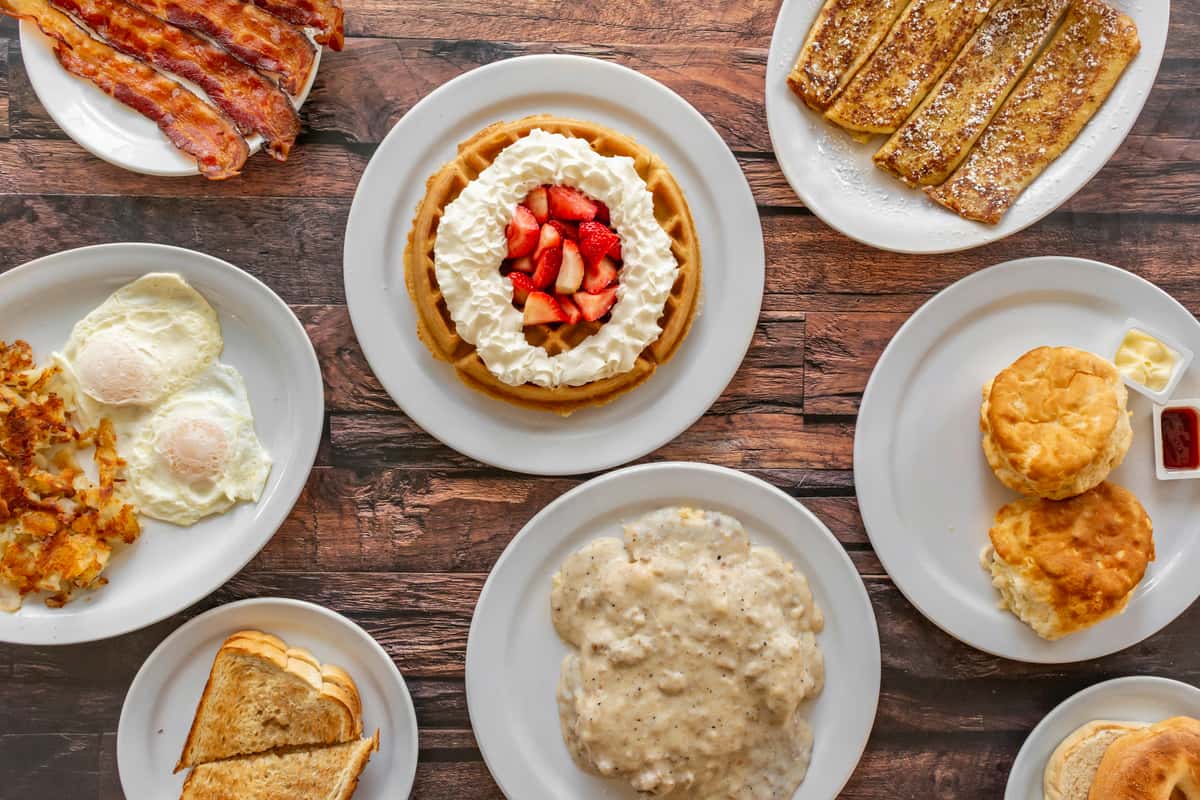 Overhead view of a hearty American breakfast spread featuring crispy bacon, sunny-side-up eggs with hash browns, buttery toast, Belgian waffle topped with whipped cream and fresh strawberries, French toast sticks, flaky biscuits with butter and jam, and a plate of biscuits smothered in creamy sausage gravy — a classic diner-style breakfast feast perfect for brunch menus.