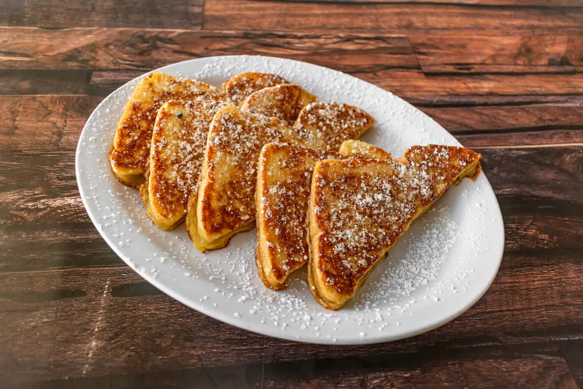 Golden-brown slices of classic French toast dusted with powdered sugar, neatly arranged on a white plate over a rustic wooden table — a comforting breakfast staple perfect for diners or brunch cafes.