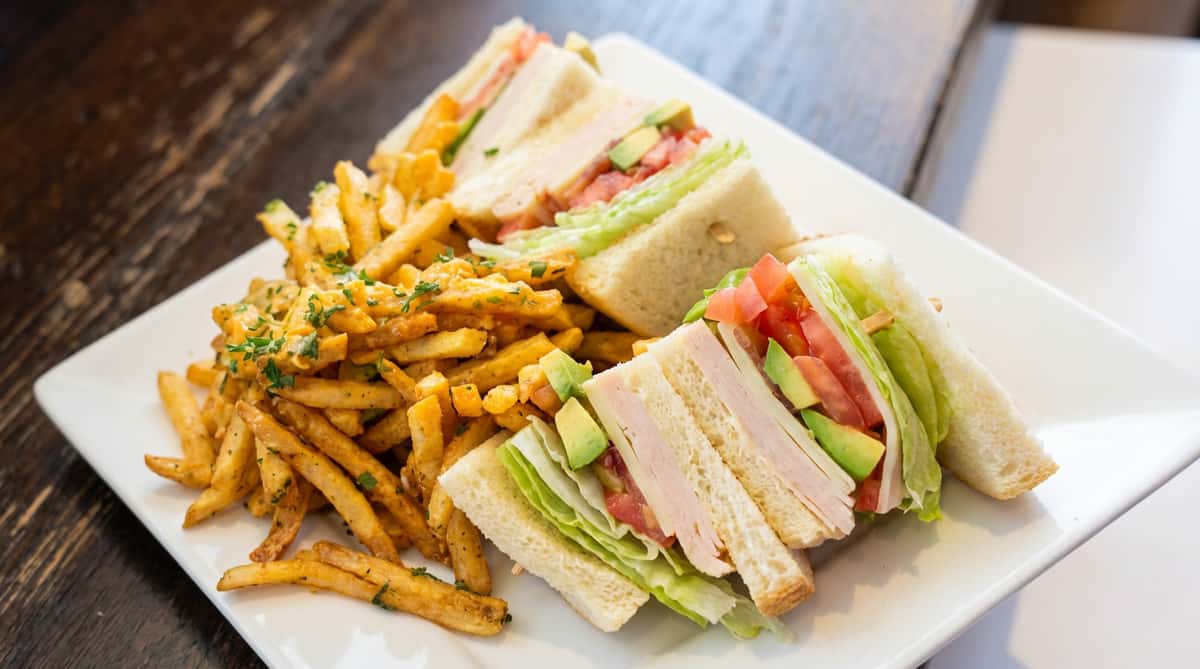 Turkey club sandwich halves with lettuce, tomato, avocado, and fries on a white rectangular plate.