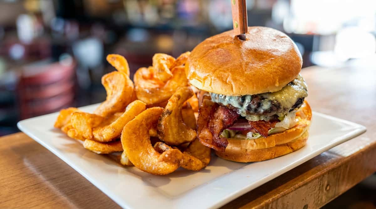 Burger with melted cheese, bacon, lettuce, tomato, and onion, served with curly fries on a white rectangular plate.