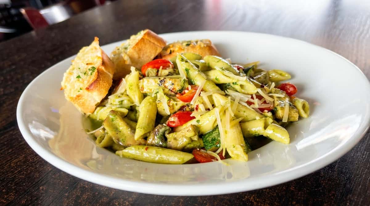 Pesto pasta with chopped tomatoes, mushrooms, and zucchini, topped with shredded Parmesan, served with garlic bread.