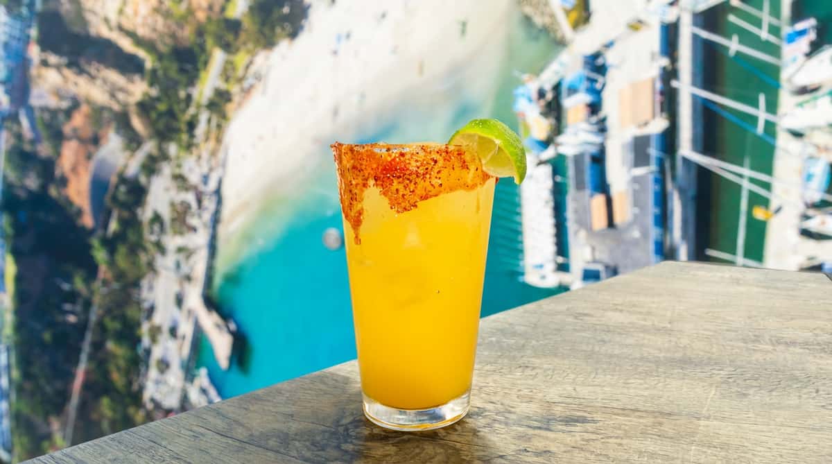 A bright orange cocktail in a tall glass with a chili-salt rim and a lime wedge garnish, set on a wooden ledge overlooking a marina with turquoise water and boats below.
