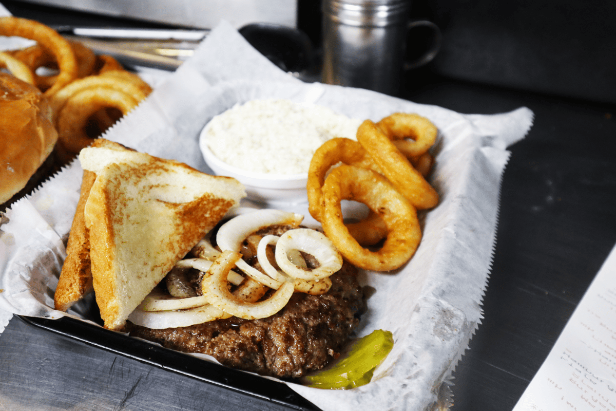 Hangry's Grill's juicy burger topped with grilled onions and toast, served with onion rings and a side of creamy sauce.