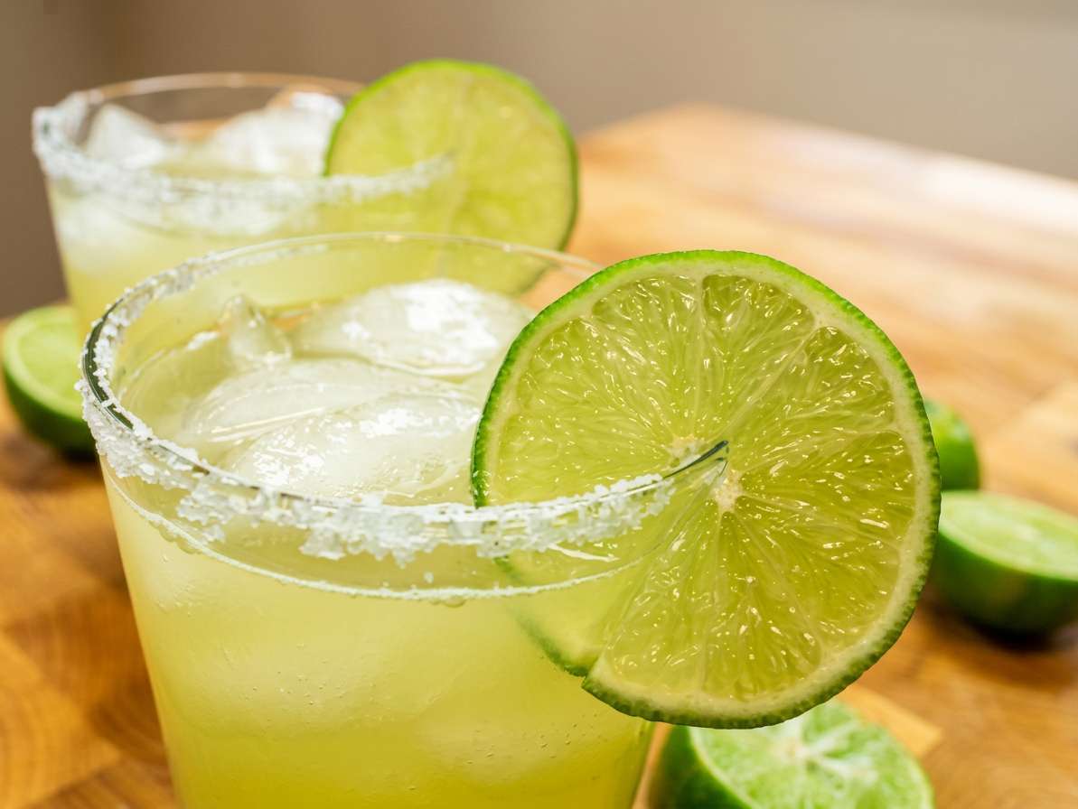 Margaritas in glasses with salted rims, lime slices, ice, and a bottle in the background.