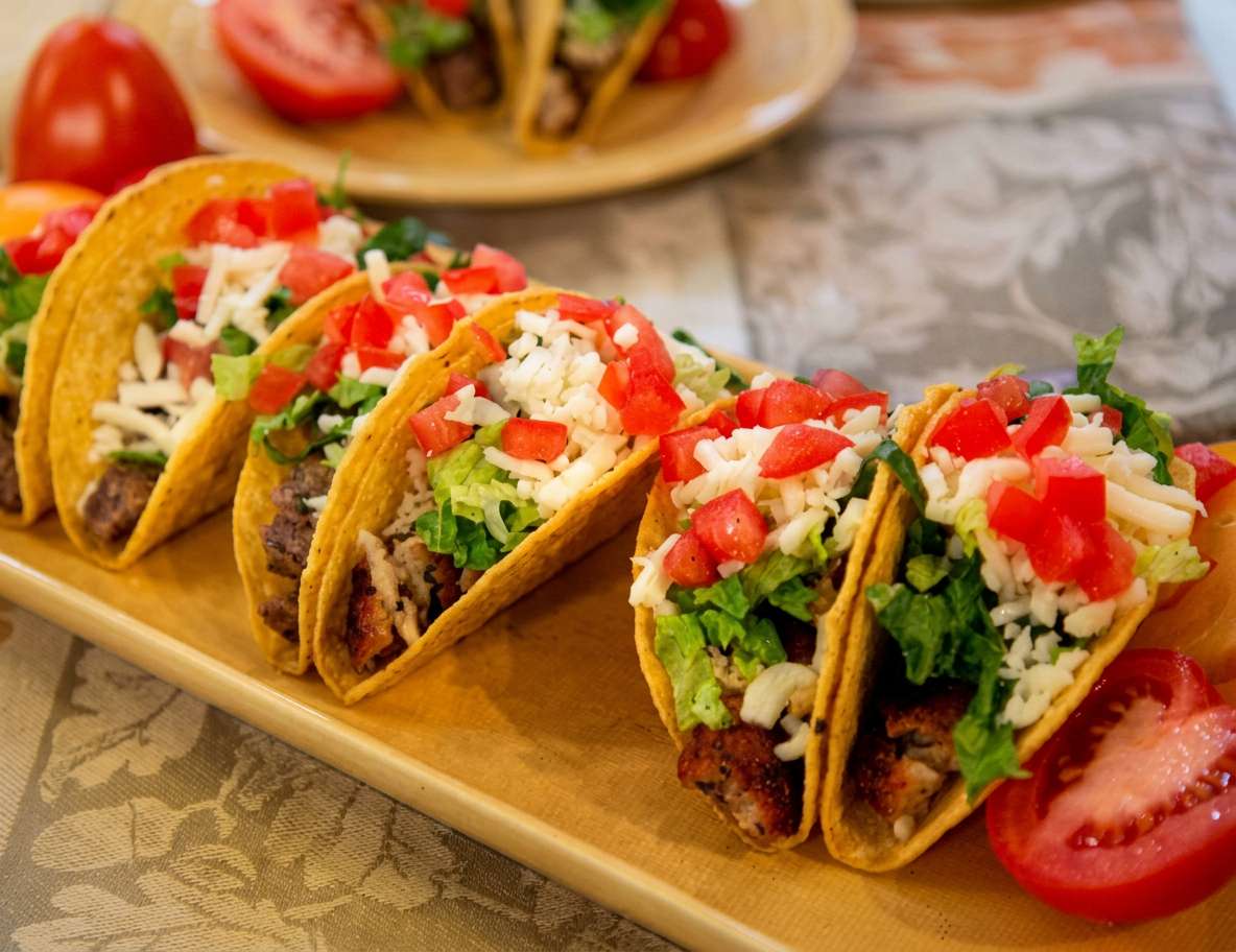 Five hard-shell tacos filled with meat, lettuce, shredded cheese, and diced tomatoes on a wooden platter.