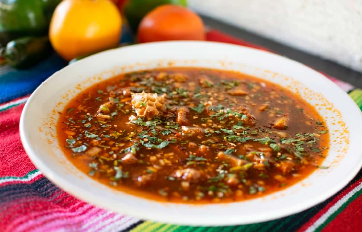 Menudo, a red chili-based soup with tripe and cilantro, in a white bowl on a colorful striped cloth.