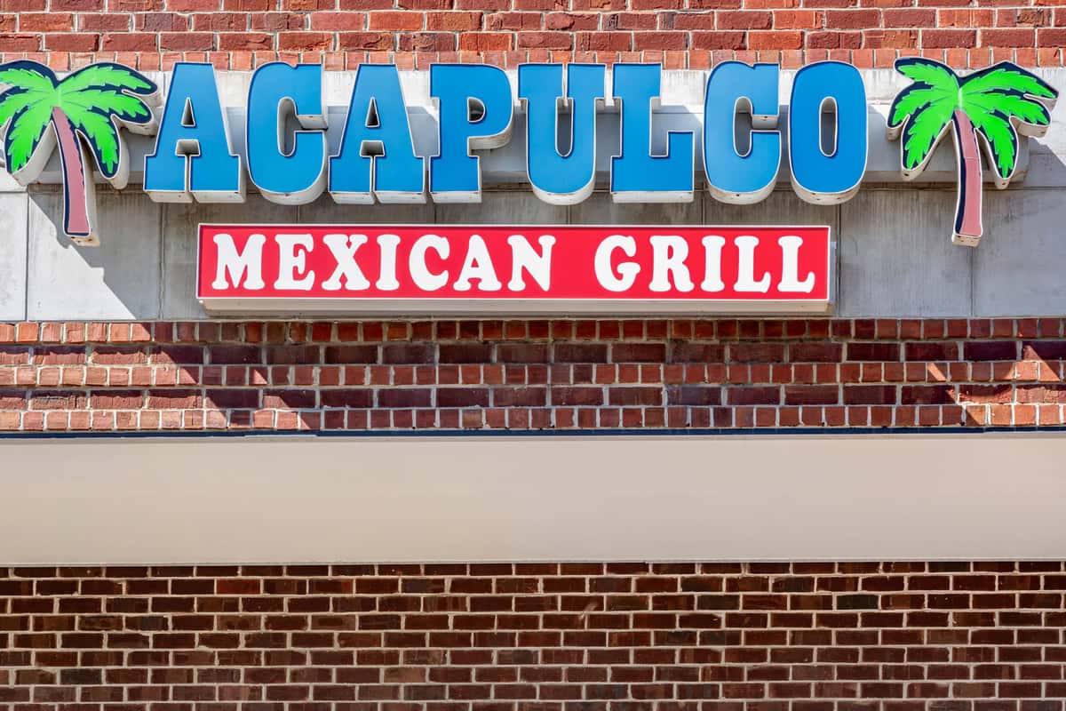Close-up of the Acapulco Mexican Grill storefront sign, featuring bold blue lettering, red and white "Mexican Grill" banner, and vibrant green palm tree icons against a red brick background.