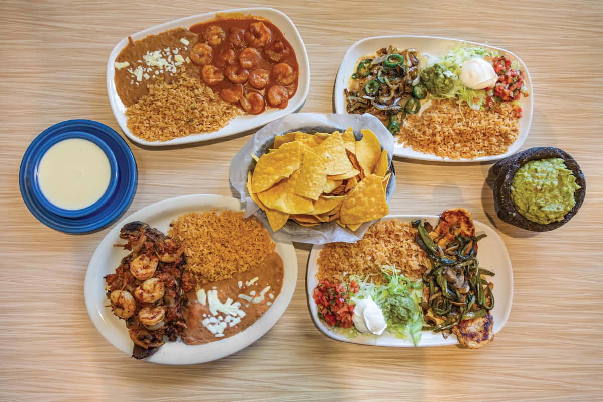 Colorful Mexican food spread on a wooden table featuring shrimp dishes with rice and refried beans, sizzling grilled vegetables, fresh pico de gallo, shredded lettuce, guacamole, sour cream, crispy tortilla chips, a stone bowl of chunky guacamole, and a side of creamy queso dip. Traditional Tex-Mex presentation ideal for family-style dining.