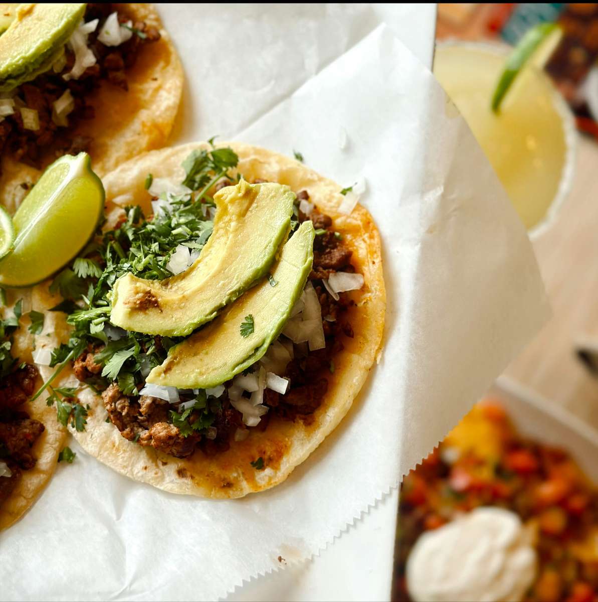Street-style carne asada tacos topped with fresh cilantro, diced onions, sliced avocado, and lime wedges, served on soft corn tortillas. A classic margarita garnished with lime is partially visible in the background, adding a refreshing touch to this vibrant Mexican dish.
