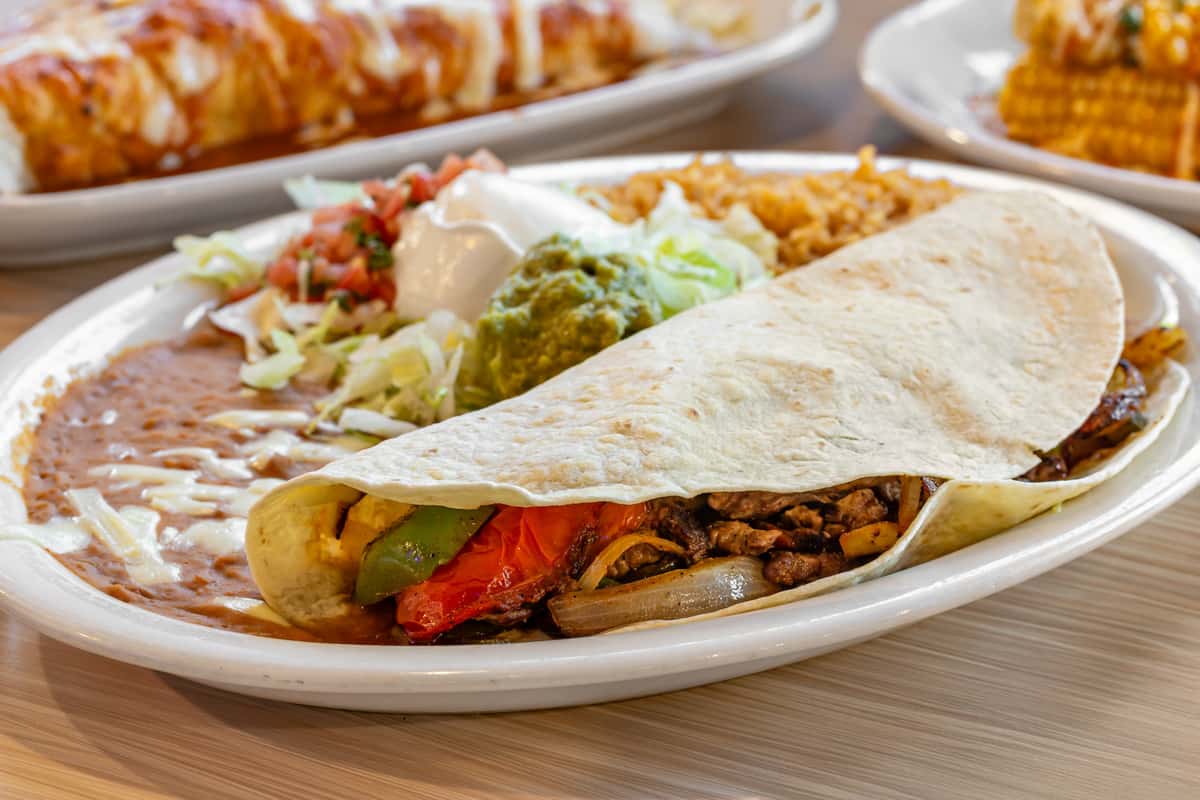 Grilled steak fajita wrapped in a soft flour tortilla with sautéed peppers and onions, served with Mexican rice, refried beans topped with melted cheese, guacamole, sour cream, shredded lettuce, and fresh pico de gallo.