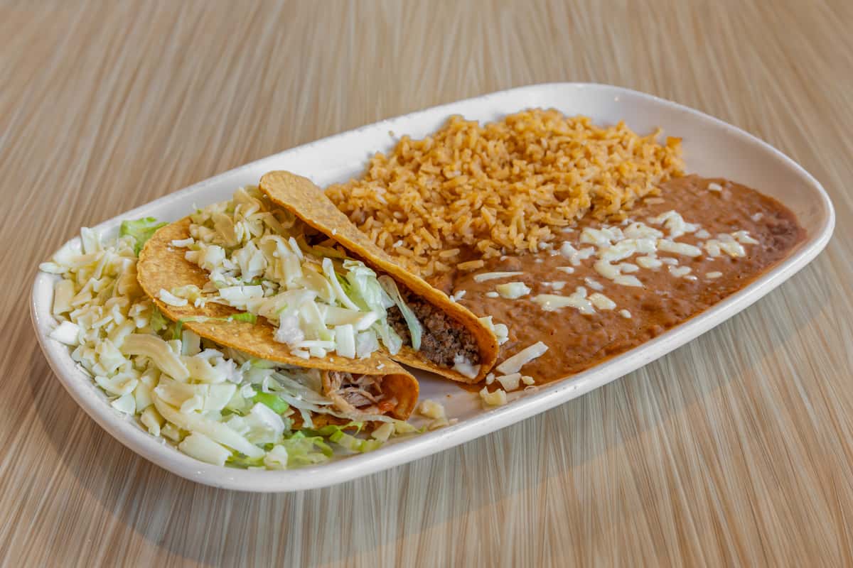 Two crispy beef tacos topped with shredded lettuce and cheese, served with Mexican rice and refried beans sprinkled with melted cheese on a white oval plate.