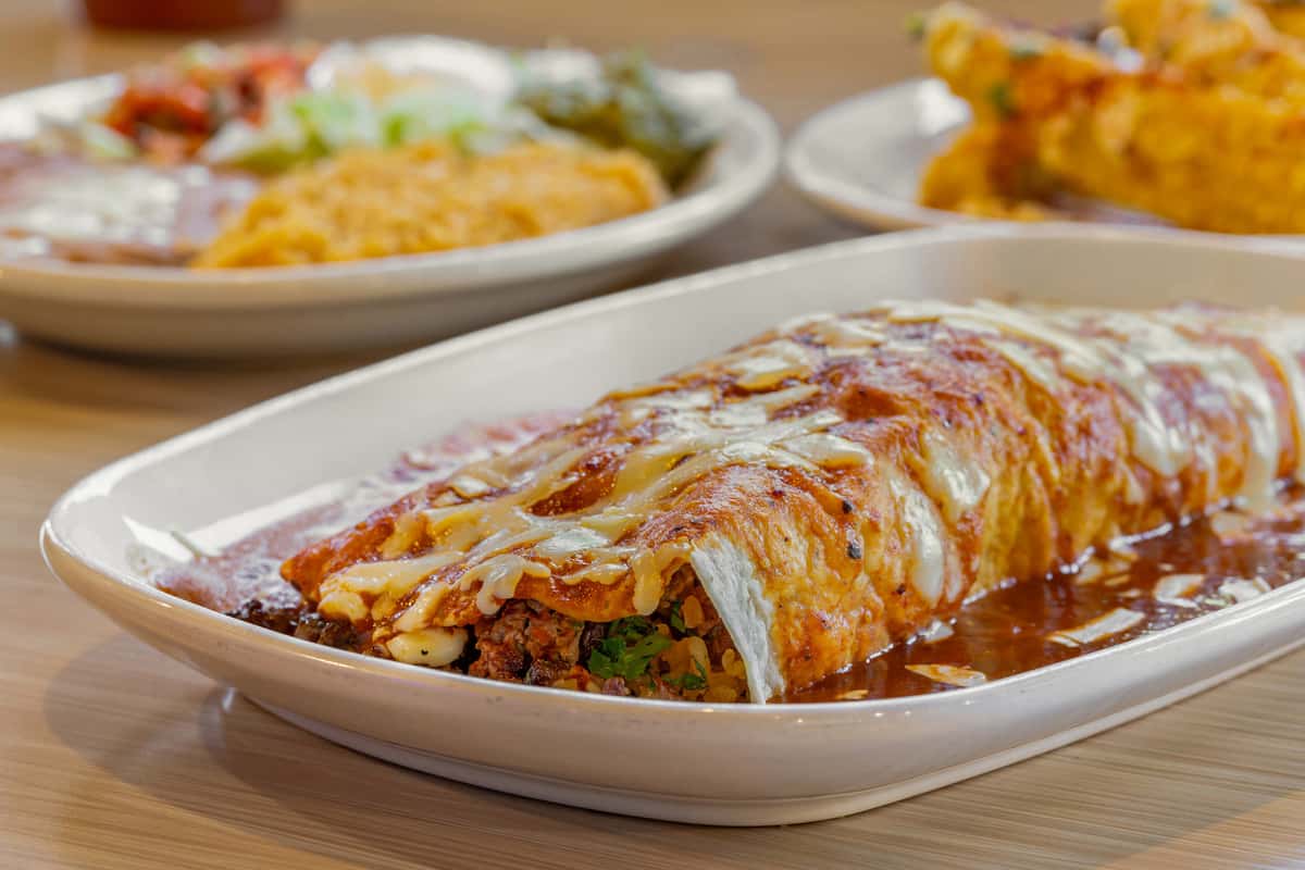 Close-up of a giant smothered burrito filled with seasoned ground beef, rice, beans, and fresh cilantro, topped with melted cheese and red enchilada sauce, served on a white plate in a vibrant Tex-Mex restaurant setting.