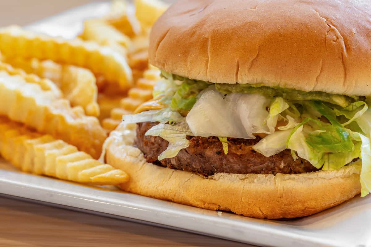 Juicy hamburger topped with shredded lettuce and onions on a toasted bun, served with crispy crinkle-cut fries on a metal tray, showcasing a classic American diner-style meal.