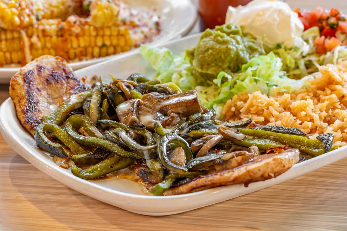 Grilled chicken breast topped with sautéed poblano peppers and mushrooms, served with Mexican rice, shredded lettuce, fresh guacamole, sour cream, and pico de gallo on a white plate at a Tex-Mex restaurant.