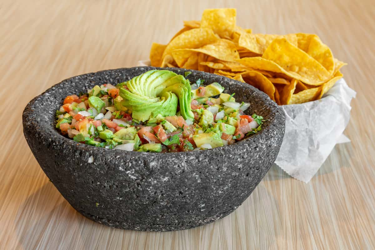 Chunky guacamole made with avocado, tomato, onion, and cilantro, served in a traditional molcajete bowl with crispy tortilla chips on the side.