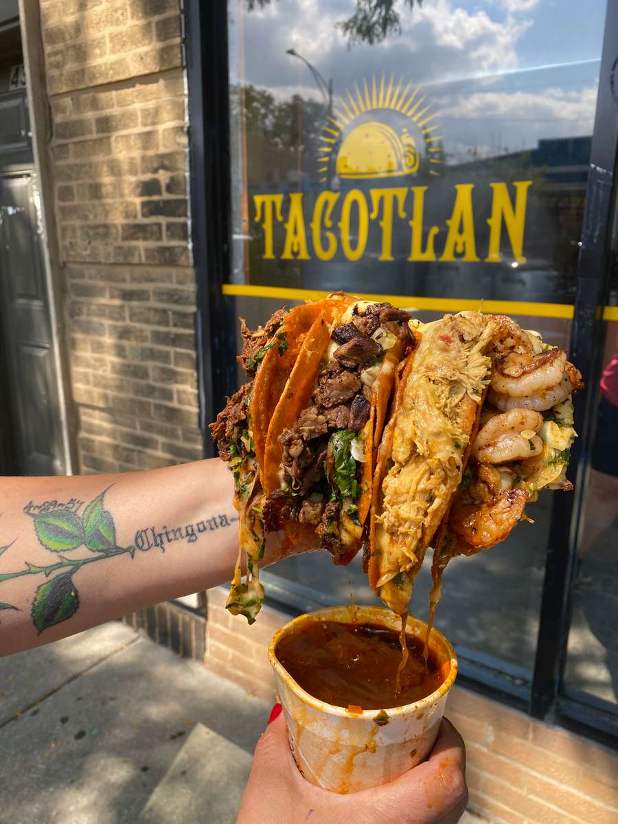 A hand holds a selection of tacos from Tacotlan, with savory fillings and a cup of sauce, showcasing delicious street food in Chicago
