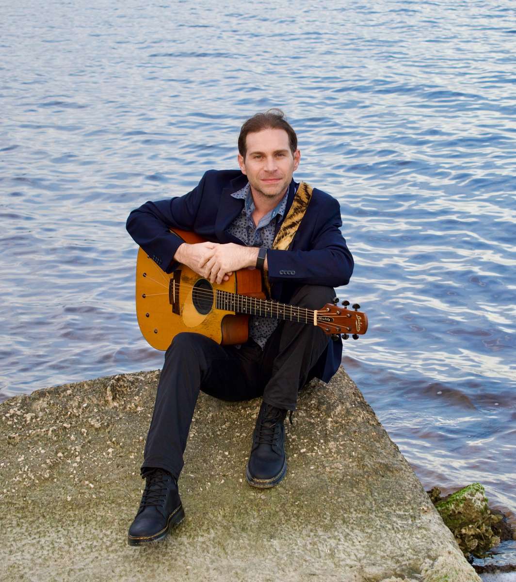 Young man in suit, sitting on edge of water holding acoustic guitar
