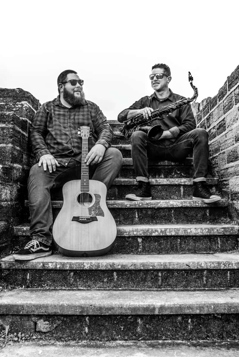 Two gentlemen sitting on brick steps, holding acoustic guitar and a wind instrument
