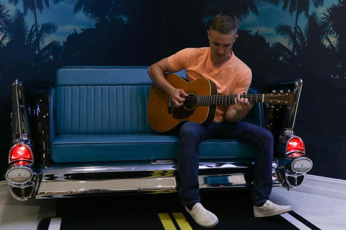 young twenties, male holding an acoustic guitar, sitting on a blue couch.