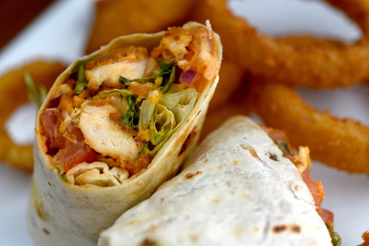 Image of a Buffalo chicken wrap sandwich with onion rings. The wrap is a tortilla containing grilled or fried chicken strips.