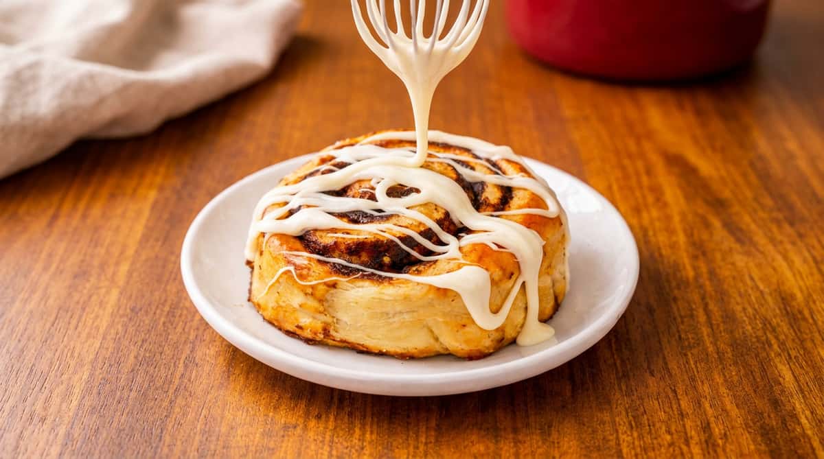 Cinnamon roll on a white plate with white icing being drizzled from a whisk.