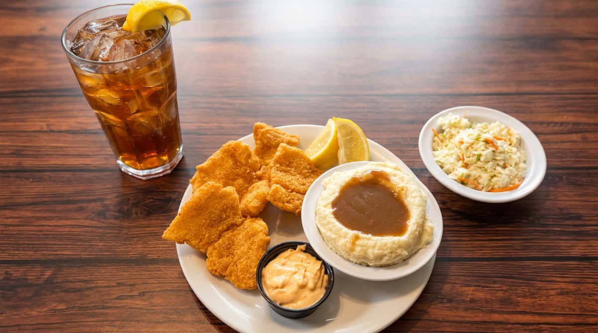Fried fish platter with mashed potatoes and gravy, coleslaw, dipping sauce, and a Coca-Cola on a wooden table.