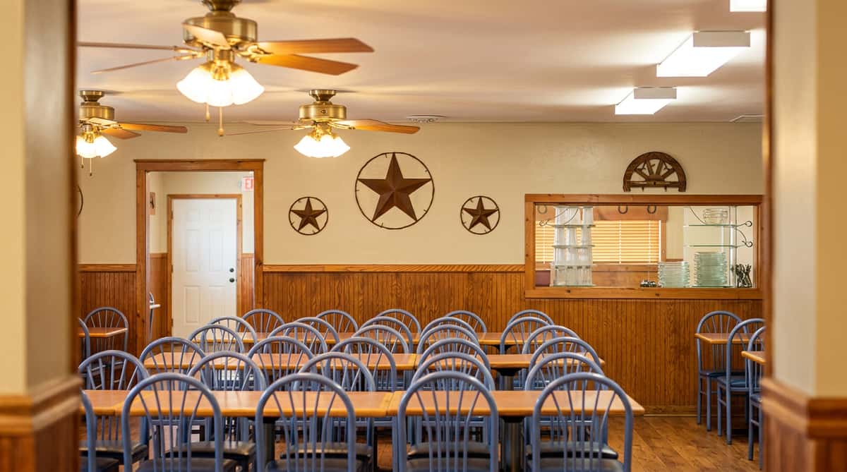 Restaurant dining room with rows of tables and chairs, wood paneling, ceiling fans, and metal star decorations.