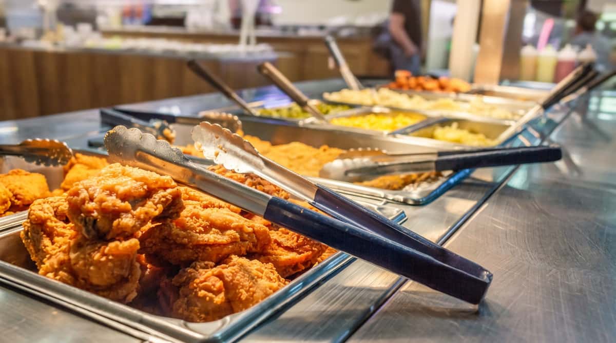 Fried chicken with tongs on a buffet line, with other food trays like mashed potatoes and macaroni in the background.