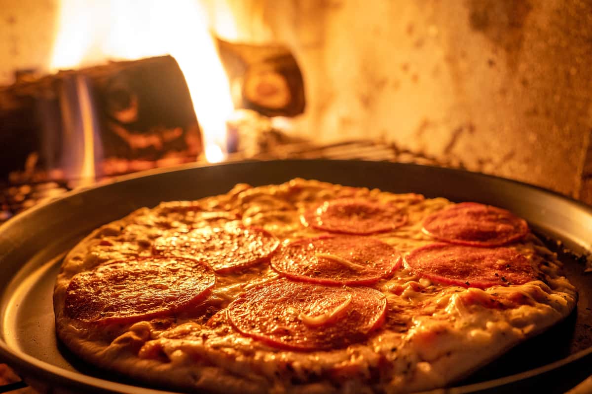 Pepperoni pizza baking in a traditional wood-fired oven with flames in the background, emphasizing the authentic smoky flavor and crispy crust — perfect for artisan or Neapolitan-style pizza branding.