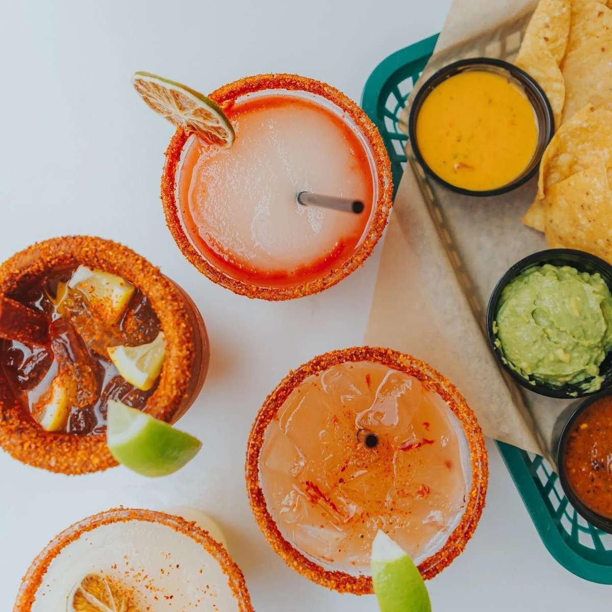 Four vibrant, chili-rimmed cocktails are presented alongside tortilla chips, guacamole, queso, and salsa from Milagro Tacos Cantina.