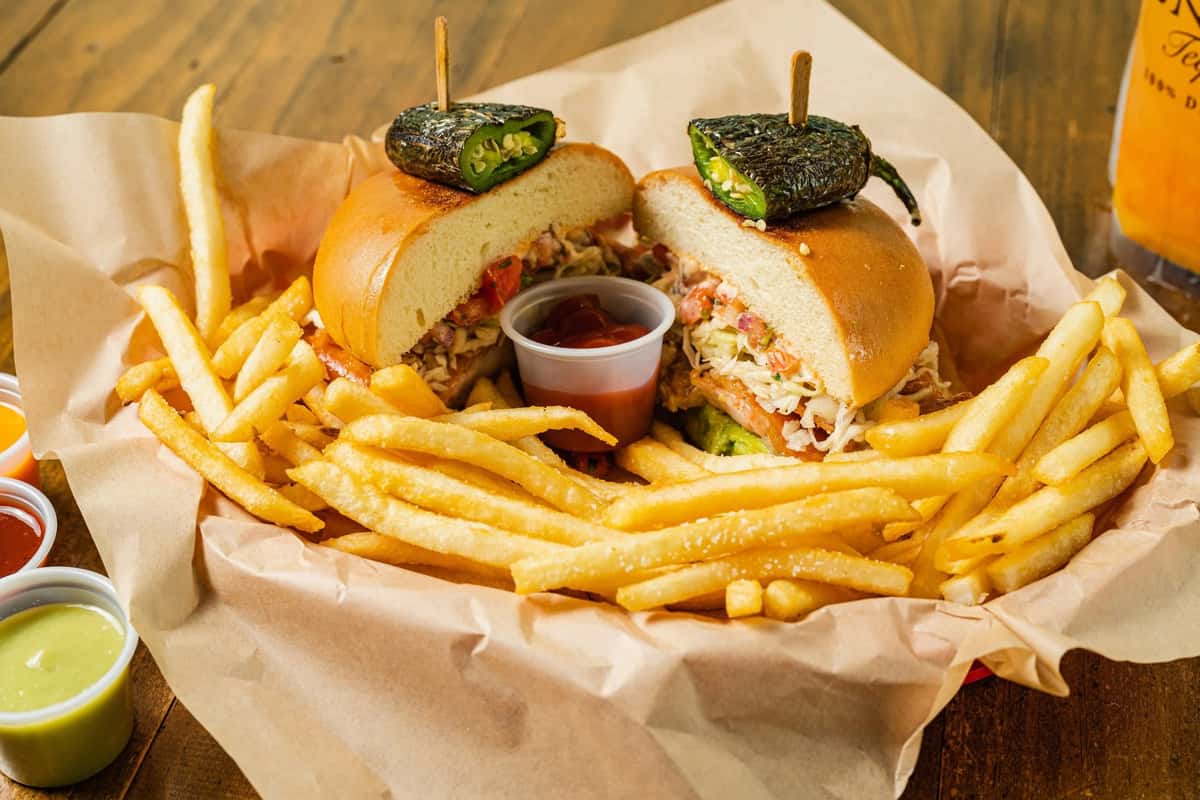 A hearty Milagro Tacos Cantina torta, cut in half and topped with roasted poblano peppers, served with golden crispy fries and colorful dipping sauces.