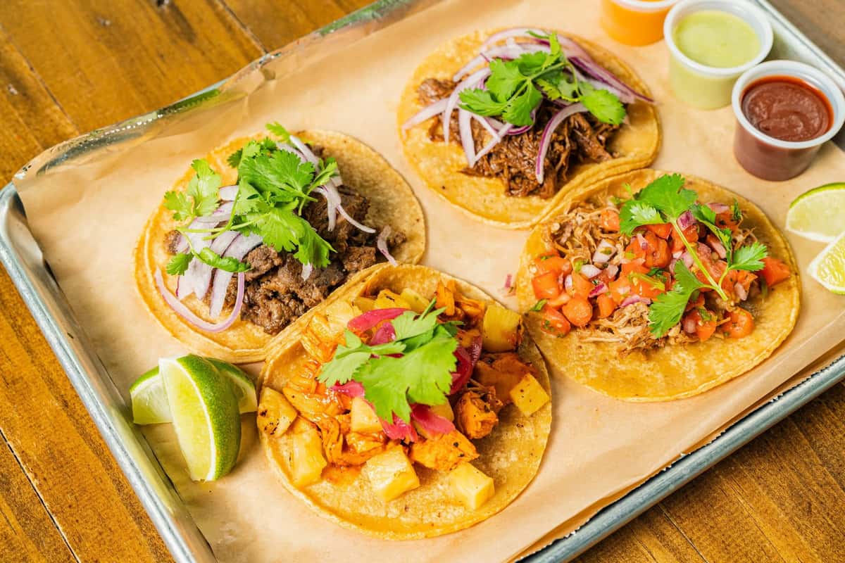 Milagro Tacos Cantina's colorful selection of tacos with various fillings, fresh toppings, lime, and sauces arranged on a metal tray.