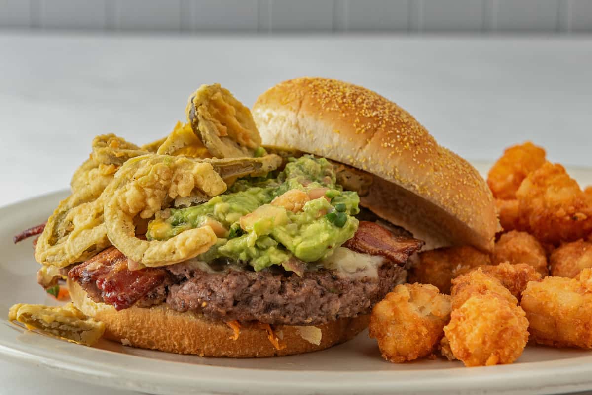 Juicy burger topped with crispy fried onions, guacamole, bacon, and jalapeños on a toasted bun, served with a side of golden tater tots — a bold twist on a classic burger.