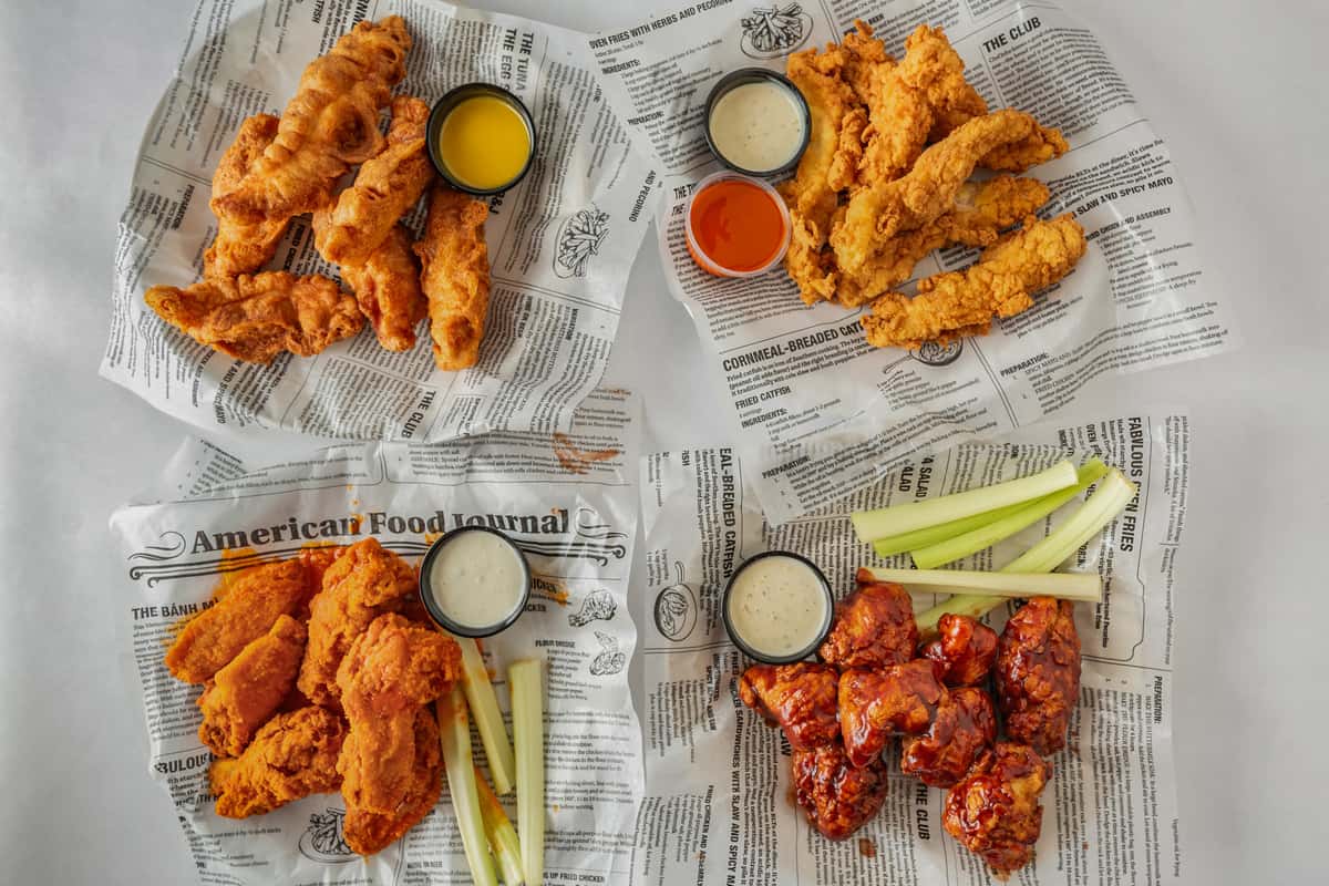 Assorted fried chicken platters served on deli-style newspaper paper, featuring crispy chicken tenders, saucy buffalo wings, celery sticks, and a variety of dipping sauces — a shareable appetizer spread perfect for game day or casual dining.