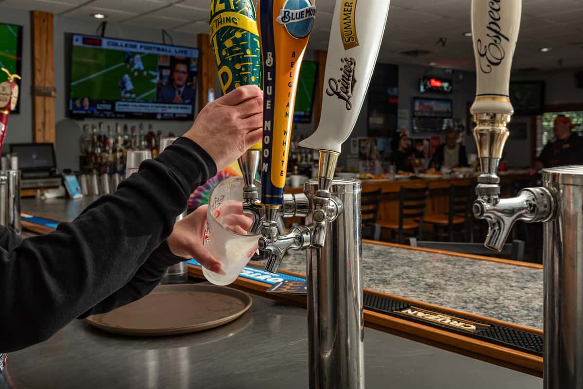 Bartender pouring a draft beer from a row of branded tap handles including Blue Moon and Leinenkugel's, set at a polished bar counter with TVs and patrons in the background — a lively sports bar scene with a focus on craft and domestic brews.