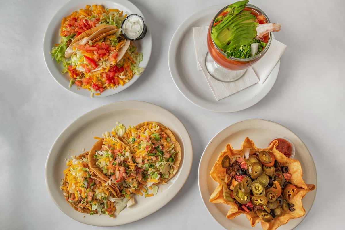 Colorful Tex-Mex combo platter featuring crunchy and soft tacos loaded with seasoned meat, shredded lettuce, diced tomatoes, and cheese, alongside a shrimp cocktail with avocado, and a crispy taco salad bowl topped with jalapeños — a vibrant spread full of fresh Mexican-inspired flavors.