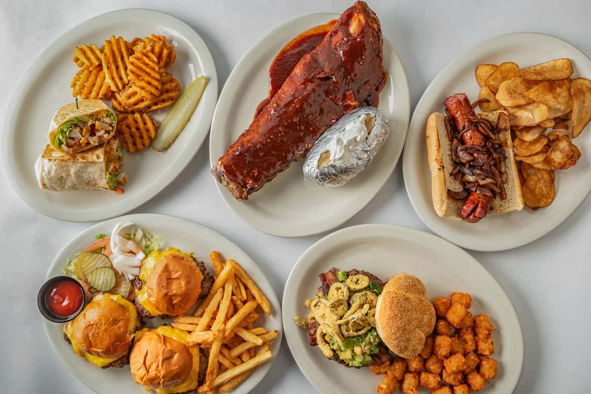 Assorted American comfort food platters featuring BBQ ribs with baked potato, a grilled sausage sandwich with chips, cheeseburger sliders with fries, a loaded burger with tater tots, a Southwest wrap with waffle fries, and more — a hearty spread perfect for casual dining or game day meals.
