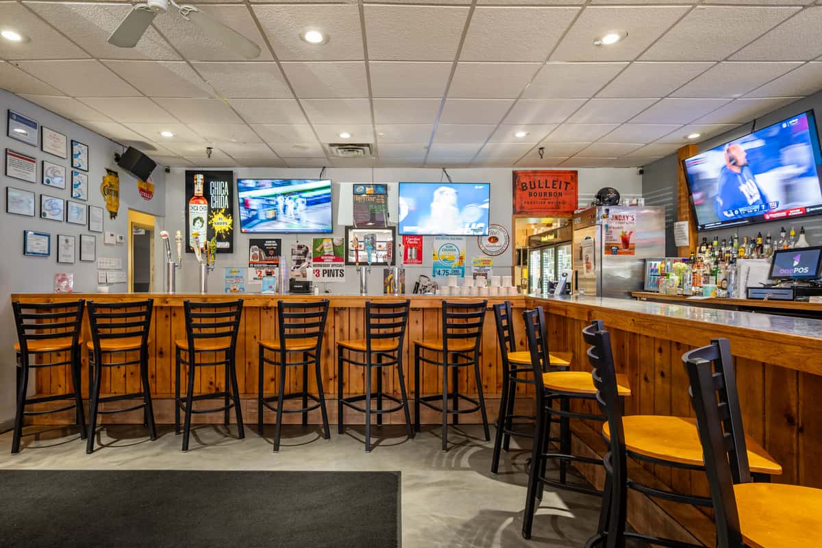 Cozy sports bar interior featuring a wooden bar counter with high-top chairs, multiple flat-screen TVs broadcasting live games, and a wide selection of beers and spirits — a lively setting for casual drinks and game day viewing.