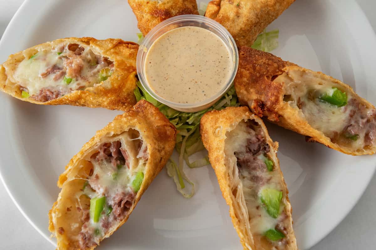 Crispy Philly cheesesteak egg rolls stuffed with shaved beef, melted cheese, onions, and green bell peppers, served with a creamy dipping sauce on a bed of shredded lettuce — a flavorful fusion appetizer perfect for sharing.
