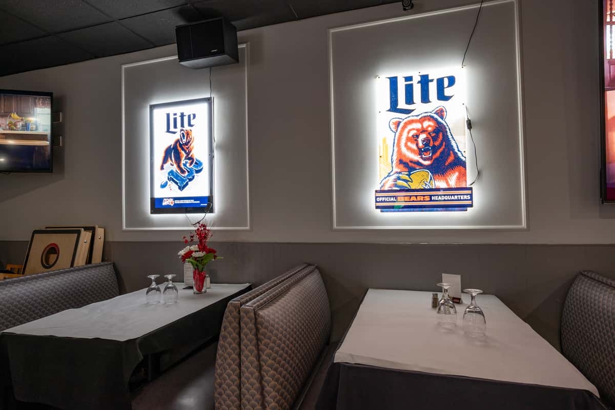 Cozy booth seating in a sports-themed restaurant, featuring Miller Lite neon signs with Chicago Bears imagery on the wall, white tablecloths with black overlays, and neatly set tables — a casual yet inviting game day dining atmosphere.