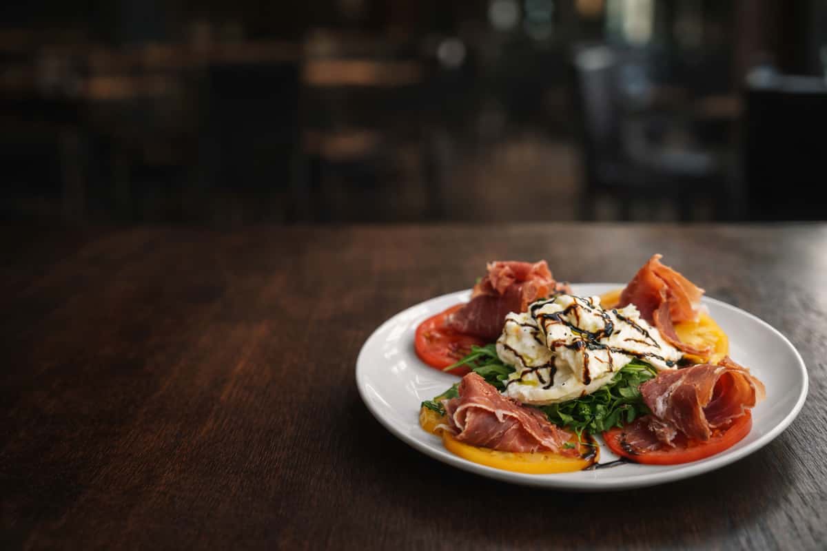 Fresh burrata with prosciutto, heirloom tomatoes, arugula, and balsamic glaze, beautifully plated on a white dish in a warm, intimate dining atmosphere.