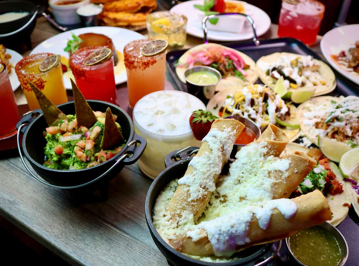 A vibrant Mexican meal spread featuring tacos, guacamole with chips, crispy flautas topped with cheese, colorful cocktails rimmed with chili, and a creamy drink garnished with a strawberry — a lively and festive dining setup.