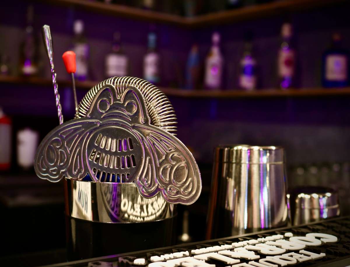 A close-up of a stylish cocktail strainer with an artistic face design, sitting on a bar counter beside a metal shaker and stirrer, with bottles of liquor softly blurred in the background — evoking a sleek and modern bar atmosphere.