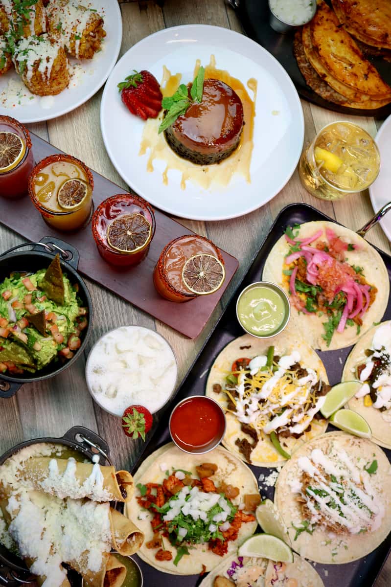 Colorful Mexican spread featuring tacos, flan dessert, elote, guacamole, and cocktails garnished with dried citrus slices.