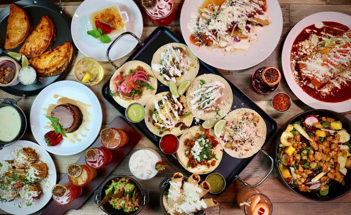 Colorful Mexican spread featuring tacos, flan dessert, elote, guacamole, and cocktails garnished with dried citrus slices.