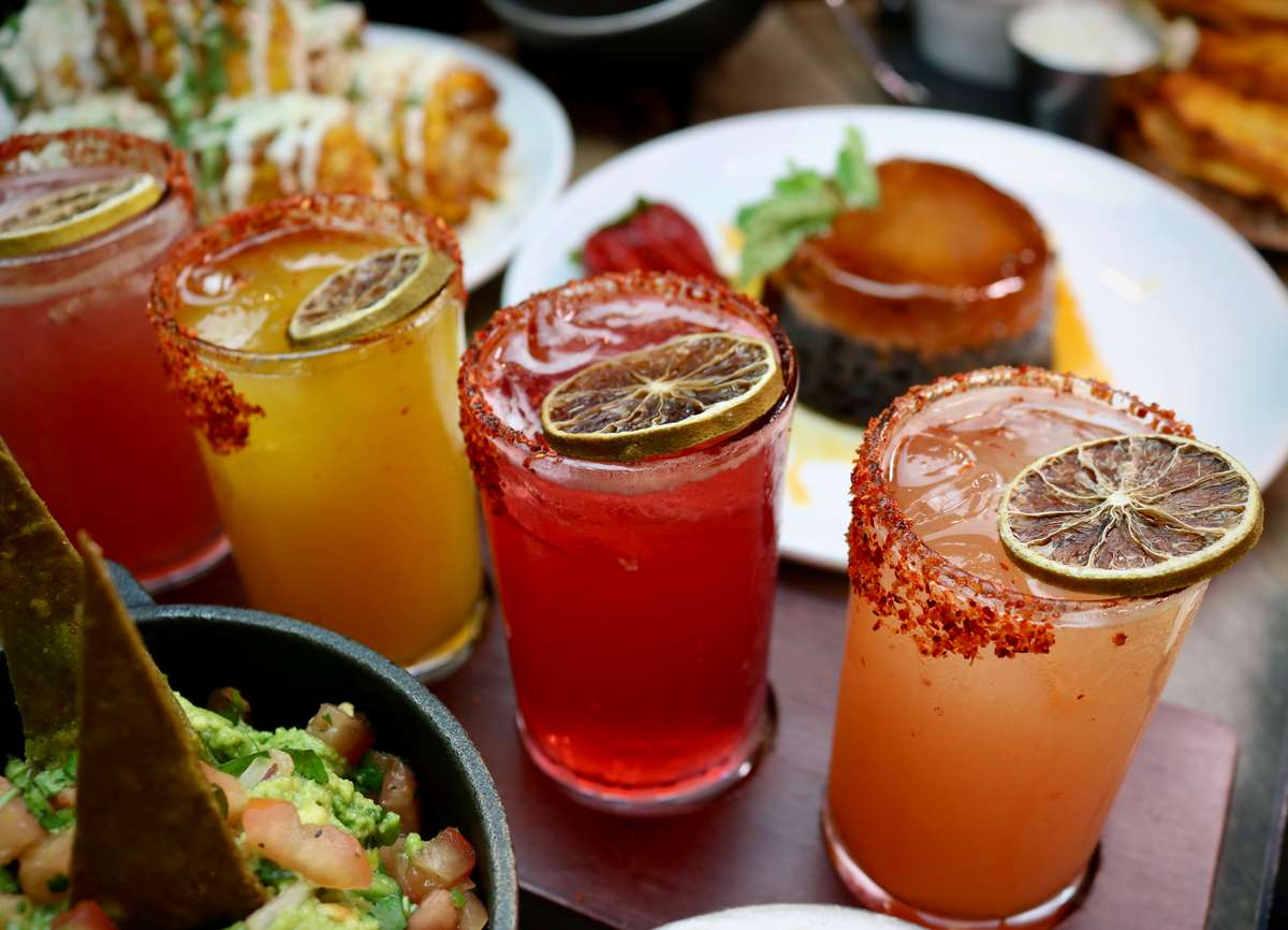 Colorful cocktails with chili-salt rims and dried lime garnishes, served alongside guacamole and a dessert flan in the background.