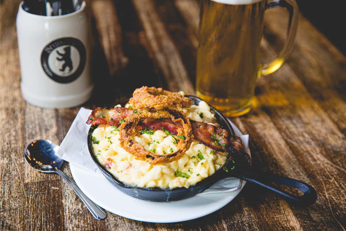 Creamy skillet mac and cheese topped with crispy bacon and fried onion rings, served alongside a traditional German beer at Berliner Pub.