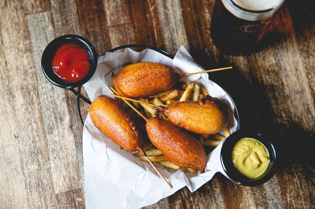 Basket of golden hand-dipped corn dogs served over seasoned fries with ketchup and mustard, paired with a German beer at Berliner Pub Beerhall.