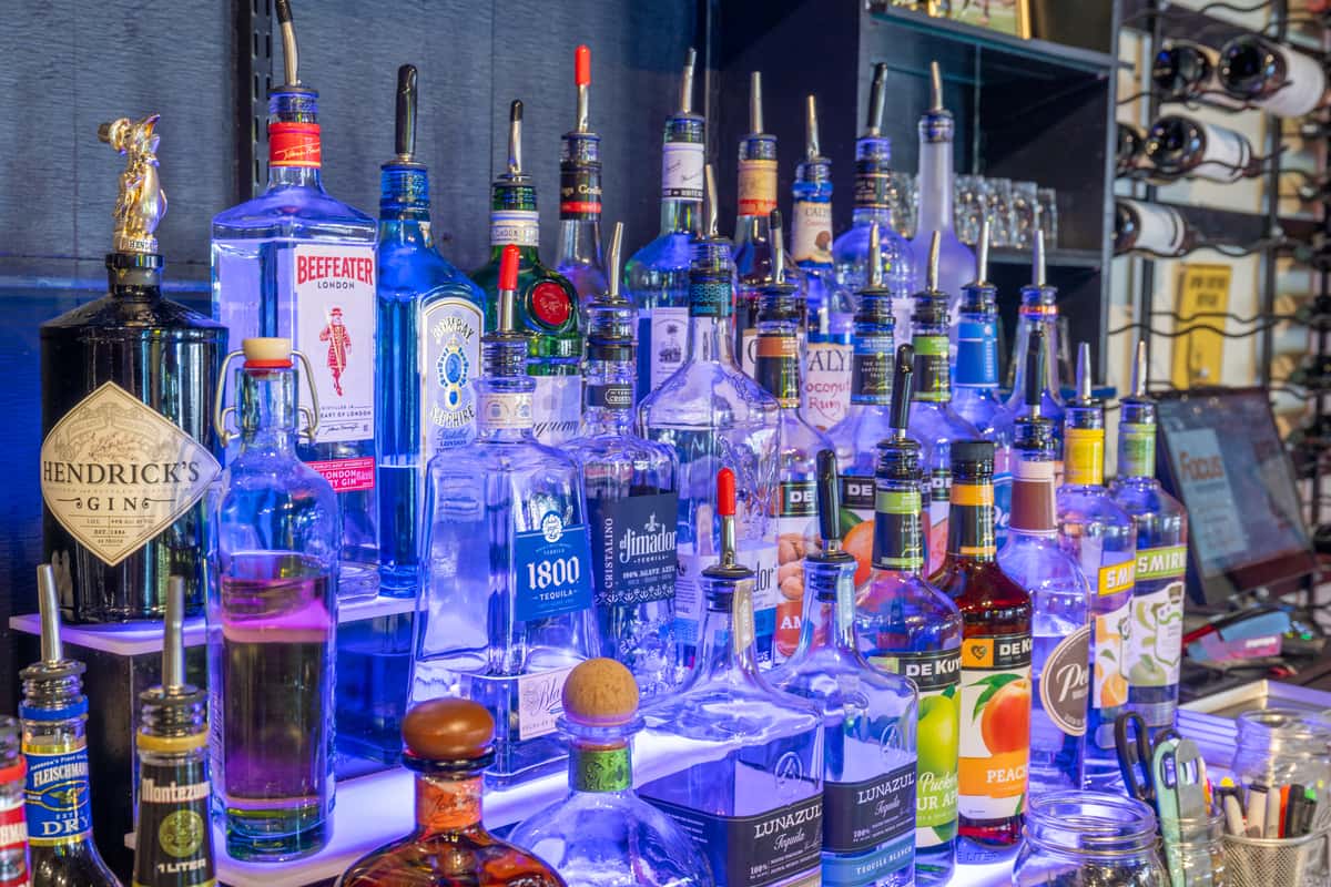 Back bar display with assorted liquor bottles arranged on illuminated shelves, glassware, and bar equipment beneath a mounted TV screen.