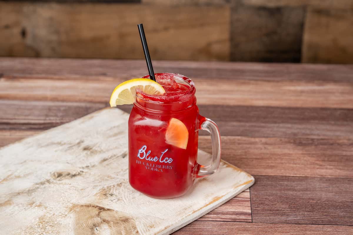 Red cocktail in mason jar with lemon wedge and black straw, Blue Ice Huckleberry Vodka logo on glass, on wood table.